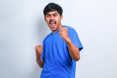 İzole edilmiş beyaz arka plan üzerinde duran sıradan tişört giyen genç yakışıklı adam mutlu ve heyecanlı bir şekilde kollarını kaldırıp, gülümseyerek ve başarı için çığlık atarak zafer hareketi yapıyor. Kutlama konsepti