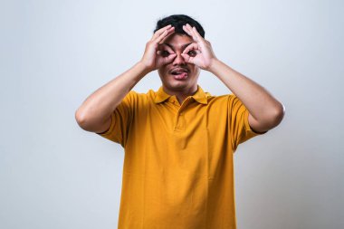 Rahat gömlekler giyen genç yakışıklı bir adam dürbün gibi jest yapıyor. Dürbün gibi dilini çıkarıyor, gözler parmaklarından bakıyor. beyaz arkaplan üzerinde çılgın ifade