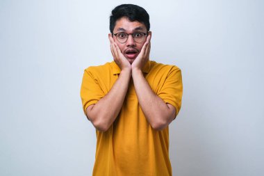 Beyaz arka plan üzerine gündelik gömlek giyen genç yakışıklı adam korkmuş ve şok olmuş, şaşırmış ve şaşkın bir yüz ifadesi var.