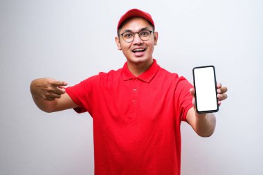 Asyalı teslimatçı, beyaz arka planda akıllı telefonu tutarken çok mutlu bir şekilde elini ve parmağını kenarı işaret ediyor.