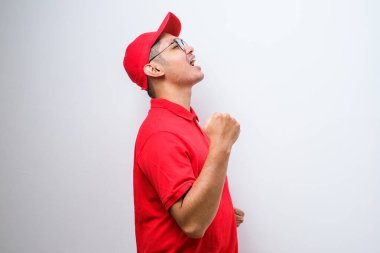 Asyalı teslimatçı, izole edilmiş kırmızı arka planda durmuş mutlu ve heyecanlı bir şekilde kollarını kaldırıp, gülümseyerek ve başarı çığlıkları atarak zafer jesti yapıyor. Kutlama konsepti.