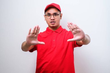 Asyalı genç teslimatçı gözlüklü ve kırmızı şapkalı, izole edilmiş beyaz arka planda, avuç içi hareketlerini durduruyor, kızgın ve hayal kırıklığına uğramış bir ifade takınıyor.