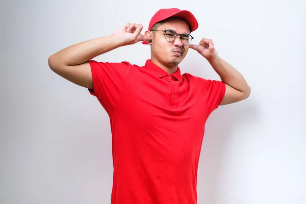 Asian delivery man wearing red uniform cover ears with hands fingers do not want to listen scream isolated on plain white background.