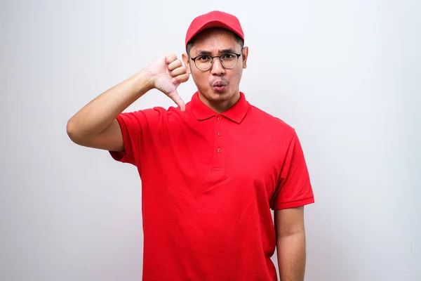 Disappointed asian delivery man in red t-shirt and cap express dislike and bad attitude, showing thumbs-down in disapproval, judging bad company service, standing white background