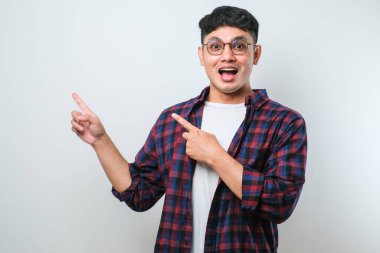 Beyaz arka plan üzerine gündelik tişört giyen yakışıklı Asyalı genç bir adam. Yüzünde kocaman bir gülümseme var. Parmağıyla kameraya bakıyor..