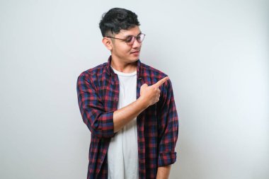 Beyaz arka plan üzerine gündelik tişört giyen yakışıklı Asyalı genç bir adam. Yüzünde kocaman bir gülümseme var. Parmağıyla kameraya bakıyor..