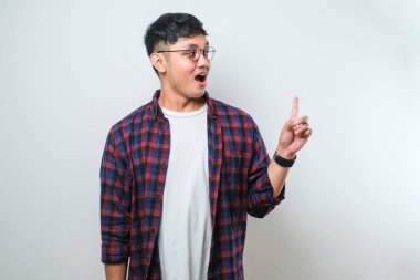 Beyaz arka plan üzerine gündelik tişört giyen yakışıklı Asyalı genç bir adam. Yüzünde kocaman bir gülümseme var. Parmağıyla kameraya bakıyor..