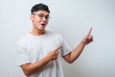 Beyaz arka plan üzerine gündelik tişört giyen yakışıklı Asyalı genç bir adam. Yüzünde kocaman bir gülümseme var. Parmağıyla kameraya bakıyor..