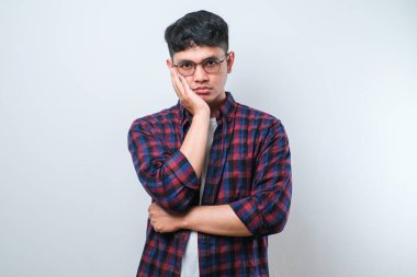 Beyaz arka plan üzerine günlük gömlek ve gözlük takan Asyalı genç adam kollarını kavuşturarak depresyondan sıkılmış ve yorgun görünüyor..