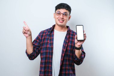 Yakışıklı Asyalı genç adam beyaz izole arka planda boş akıllı telefon ekranı gösteriyor. Çok mutlu bir şekilde elini ve parmağını kenarı işaret ediyor.