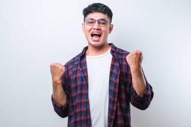 Asyalı genç bir adam, izole edilmiş beyaz arka planda dikilmiş mutlu ve heyecanlı bir şekilde kollarını kaldırıp, gülümseyerek ve başarı çığlıkları atarak zafer jesti yapıyor. Kutlama konsepti