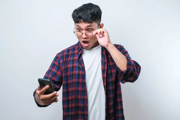 Asian young man wearing casual shirt and glasses looks surprised at the good news he received from his smartphone over white background