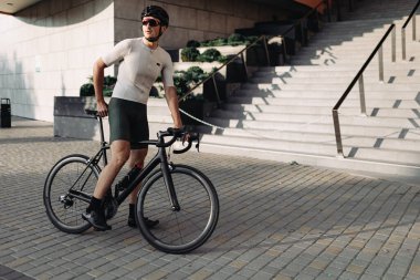 Aktif giyimli, kasklı ve gözlüklü genç adam şehir merkezinde antrenman yaptıktan sonra siyah bisikletin üzerinde oturuyor. İnsan kavramı, spor ve eğlence..