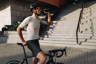 Yorgun bisikletçinin kentsel ortamlardaki yoğun egzersizden sonra suyla canlanmasının yan görüntüsü. Spor kıyafetleri, miğfer ve gözlük takan kaslı adam..