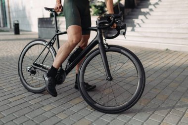 Açık havada siyah bisiklette oturan spor elbiseli aktif bir adam. Kaslı bacaklara ve tekerleklere odaklan. Kentsel alanda çalıştıktan sonra motosikletçi mola aldı.