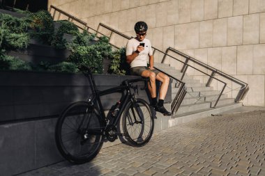 Spor elbiseli, kasklı ve gözlüklü aktif bir genç dışarıda bisiklet sürdükten sonra cep telefonunu elinde tutuyor. Malzemeler, insanlar ve aktif yaşam tarzları konsepti.