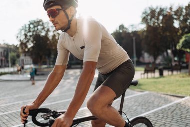 Sıcak yaz gününde aktif giyimli, kasklı ve gözlüklü bir adam şehir caddesinde bisiklet sürüyor. Beyaz tenli sporcu temiz havada en sevdiği hobiyi yapıyor..