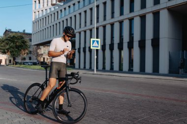 Kasklı ve gözlüklü profesyonel bisikletçi şehir caddesinde spor bisikletli ve cep telefonuyla internette sörf yapıyor. Arka plandaki modern bina.