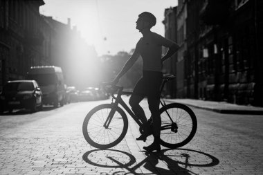 Spor kıyafetleri, miğfer ve gözlüklü yorgun sporcunun antrenman sırasında bisiklet ile şehir caddesinde duruşu. Bisiklet kavramı, insanlar ve aktif hobi. Tek renkli fotoğraf..