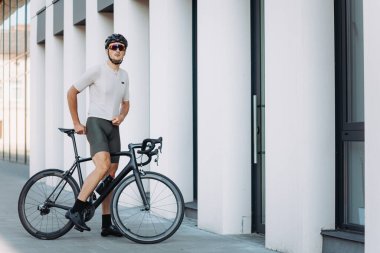 Modern binanın yanında bisiklete binen kaslı adam antrenman sırasında mola veriyor. Spor kıyafetleri giyen, kasklı ve aynalı gözlüklü profesyonel bir bisikletçi..
