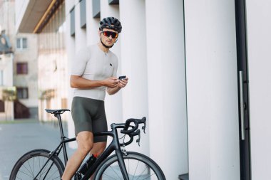 Spor elbiseli, kasklı ve gözlüklü beyaz adam dışarıda bisiklet sürerken mesafeyi ve hızı kontrol etmek için modern takip cihazı kullanıyor. Teknoloji, insanlar ve eğitim kavramı.