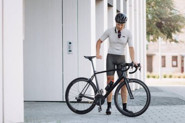 Aktif giyimli ve kasklı kaslı adam siyah profesyonel bisikletin yanında dururken aşağıya bakıyor. Kentsel ortamlarda eğitim sırasında dinlenen profesyonel bisikletçi.