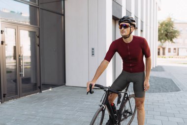 Kasklı ve gözlüklü sporcu, spor yaparken bisikletle şehir caddesinde dikiliyor. Temiz hava için kardiyo eğitimi.