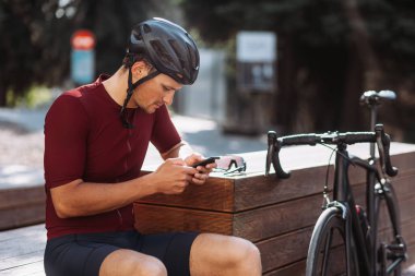 Kasklı profesyonel bir bisikletçi, ahşap bankta oturuyor ve modern akıllı telefon kullanıyor. Siyah spor bisikleti yanında duruyor. Eğlence, insanlar ve aletler konsepti.