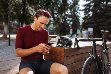 Aktif giyimli yorgun bir adam dışarıda siyah bisikletinin yanında oturuyor ve modern akıllı telefonuyla internette sörf yapıyor. Antrenmandan sonra elinde cep telefonuyla dinlenen bisikletçi..