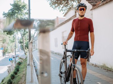 Spor elbiseli, kasklı ve gözlüklü profesyonel bisikletçi şehir caddesinde siyah bisikletin yanında duruyor. Beyaz, kaslı bacaklı bir adam dışarıda idman yaptıktan sonra dinleniyor..