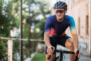 Şehir caddesinde siyah bir bisikletin üzerinde otururken yan bakan mutlu bisikletçi. Renkli spor kıyafetler, miğfer ve gözlük takan beyaz bir adam..