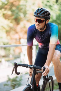 Spor kıyafetleri, kask ve gözlük giyen profesyonel bir bisikletçi açık havada bisiklet sürdükten sonra bisiklette dinleniyor. Beyaz adam temiz hava eğitiminin keyfini çıkarıyor..