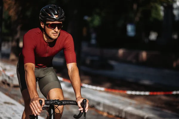 Healthy muscular guy in sport clothes, helmet and glasses cycling on black bike on empty city street. Concept of people, workout and active lifestyles.