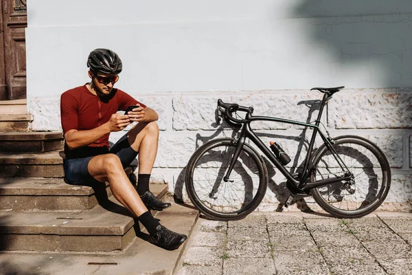Kasklı ve gözlüklü beyaz bisikletçi siyah motorsikletin yanında oturuyor ve modern akıllı telefon kullanıyor. Antrenmandan sonra dinlenme teknolojisi.