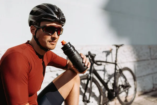 Kasklı ve bardaklı beyaz bisikletçi dışarıda antrenman yaparken spor şişesinden su içiyor. Spor etkinliğinden sonra ferahlatıcı.