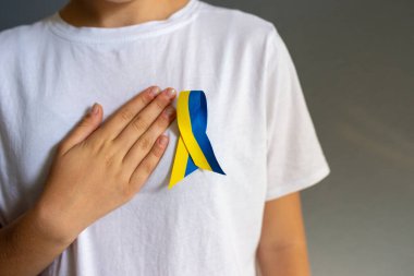 Genç bir kadın tarafından giyilen beyaz tişörtte sarı ve mavi kurdele. Elleri kalbinde.