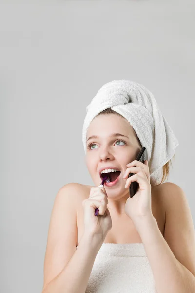 驚く美しい若い女性は、頭の上の白いタオルで彼女の歯をブラッシングしながら携帯電話で話しています。 — ストック写真