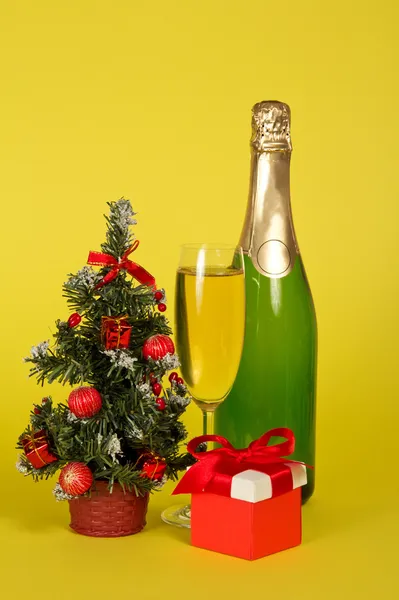 Champagne, small fir-tree in a pot with toys and gifts on a yellow background — Stock Photo, Image