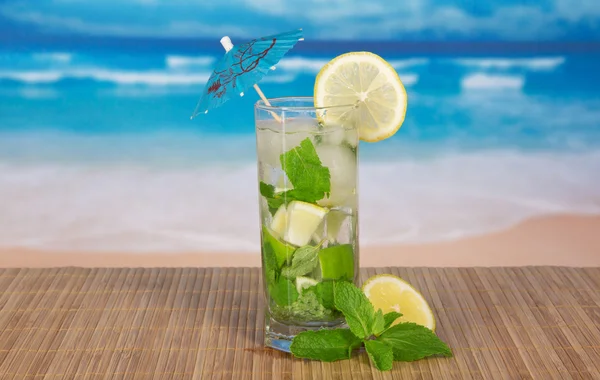 Coquetel verde de álcool com um guarda-chuva, um limão e hortelã, em um pano de bambu contra o mar — Fotografia de Stock