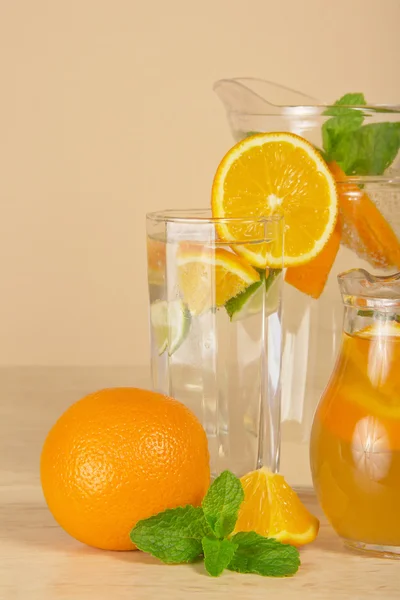 Jugs with drinks and a glass, oranges and spearmint on a beige table — Stock Photo, Image