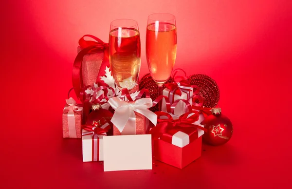 Wine glasses with champagne, gift boxes, Christmas spheres, snowflake and blank card on a red background — Stock Photo, Image