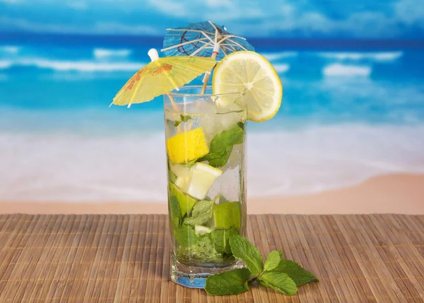 Green cocktail of alcohol with an umbrella, a lemon and spearmint, on a bamboo cloth against the sea — Stock Photo, Image