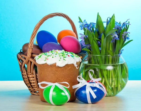 Easter cake, basket with eggs, a vase with the flowers on a table, on a blue background — Stock Photo, Image