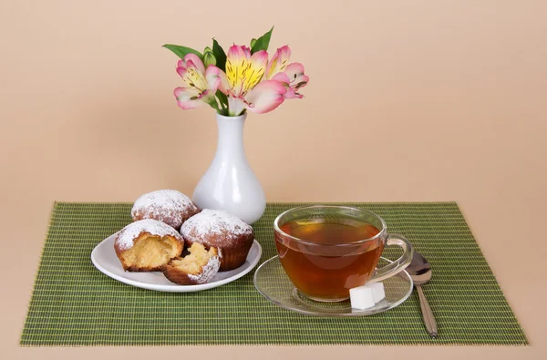 Tea, cupcakes and vase with the flowers on a bamboo cloth, on a beige background — Stock Photo, Image