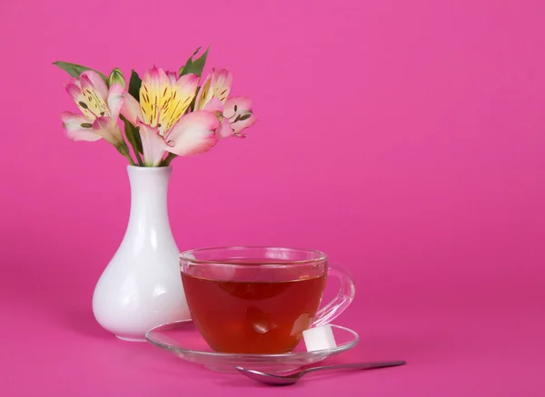 Cup of tea and sugar on a saucer, a teaspoon, vase with the flowers, on a pink background — Stock Photo, Image