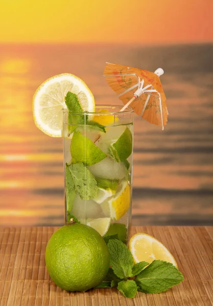Glas mit einem Mojito, einer saftigen Limette und einem Minzblatt auf einem Bambustuch gegen den Sonnenuntergang — Stockfoto