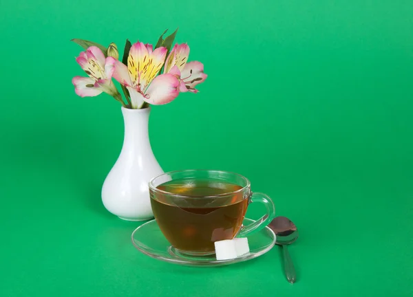 Cup of tea and sugar on a saucer, a teaspoon, vase with the flowers, on a green background — Stock Photo, Image