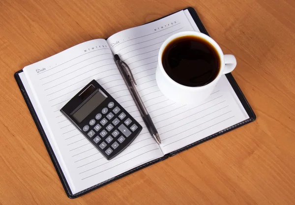 Bloco de notas aberto, a maçaneta, a calculadora e a xícara de café em uma mesa — Fotografia de Stock