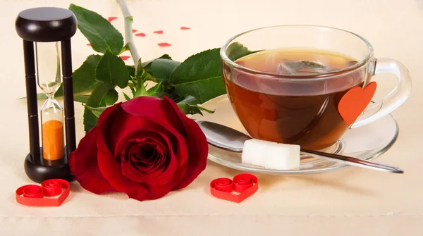 Hot tea, red rose and hourglasses on a cloth decorated with hearts — Stock Photo, Image