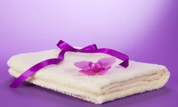 The fluffy, terry towel decorated with a flower of an orchid and a ribbon on a violet background — Stock Photo, Image